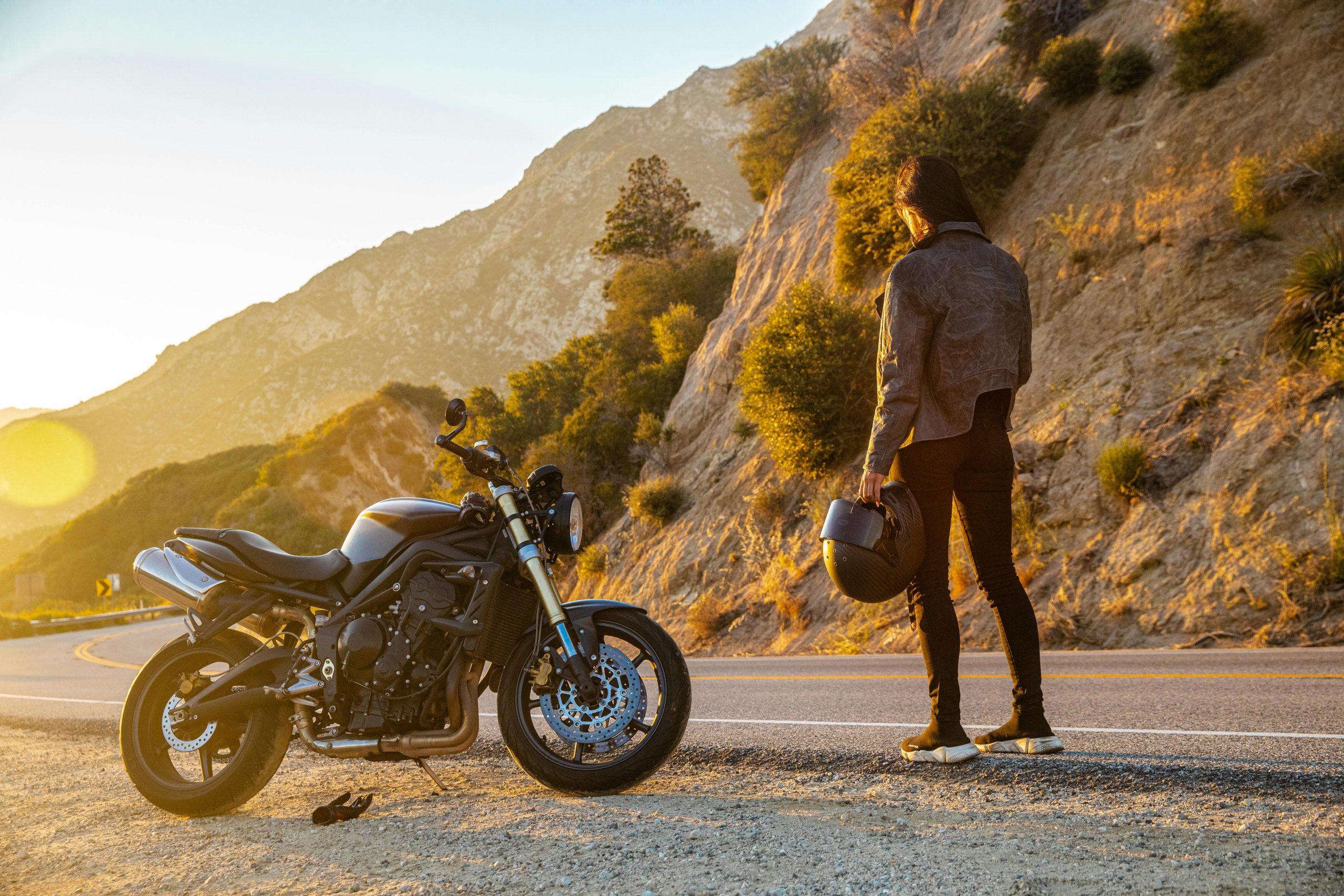 A motorcyclist stands by their bike on a mountain road at sunset, enjoying the view.