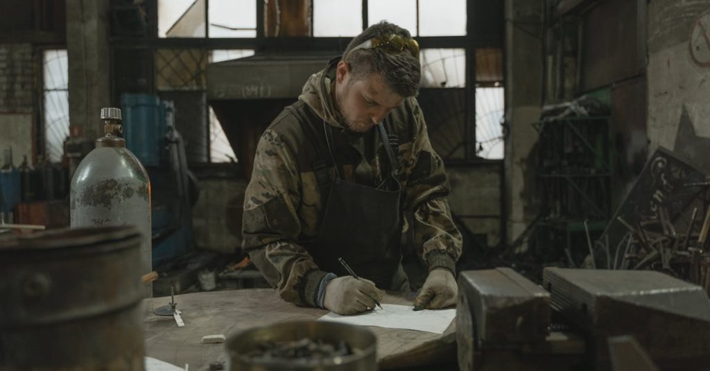 A metalworker in camouflage jacket sketches designs in an industrial workshop.